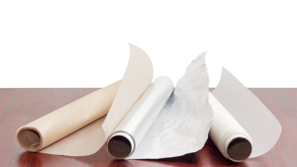 Two rolls of the various parchment paper and one roll of the aluminum foil for household use on a dark red wooden table on a white background