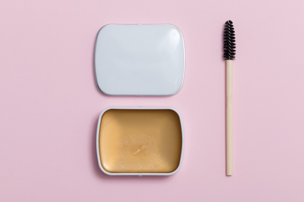 A soap brow product and spoolie brush on a pink background