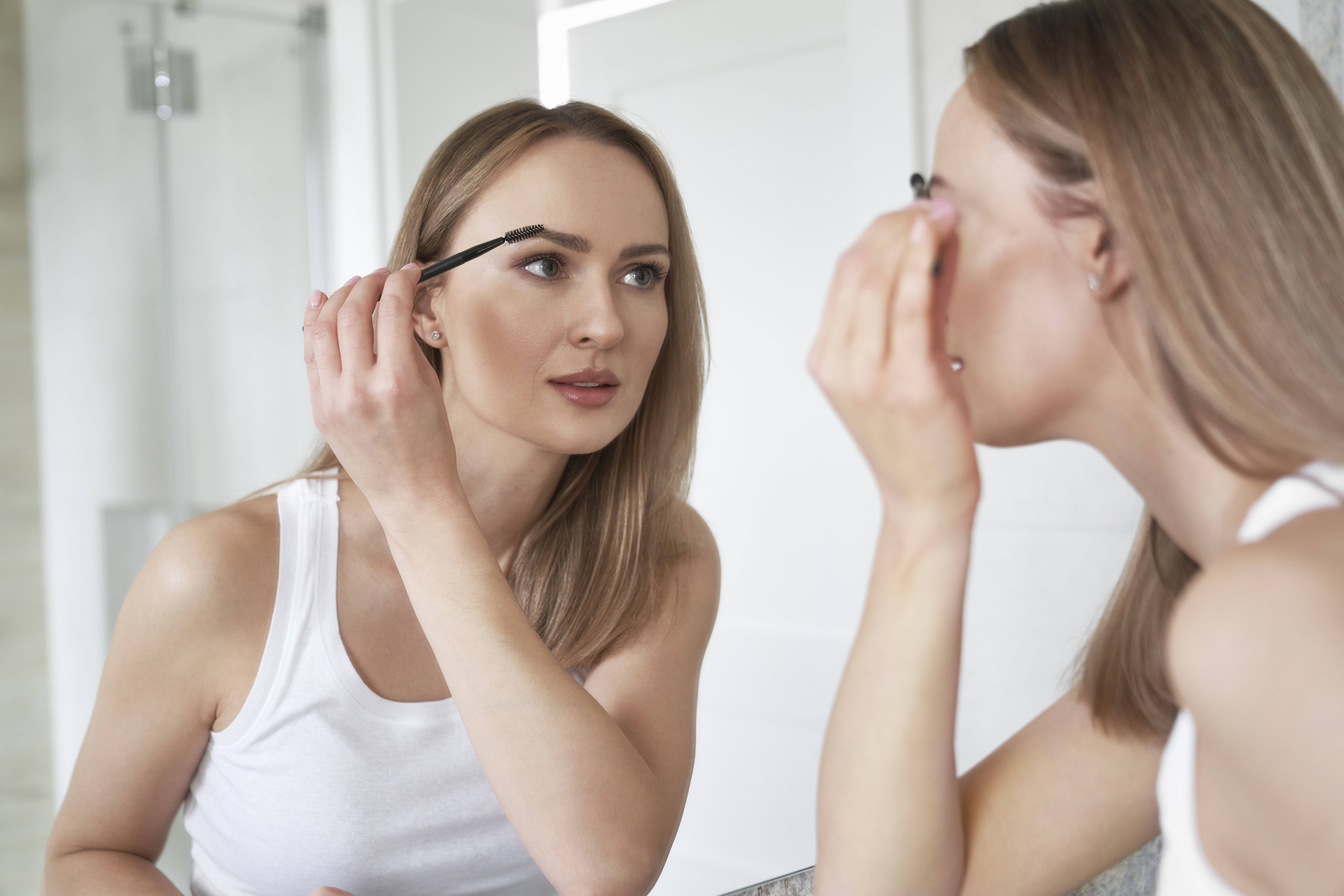 A woman looking in the mirror and brushing her eyebrows using a spoolie brush