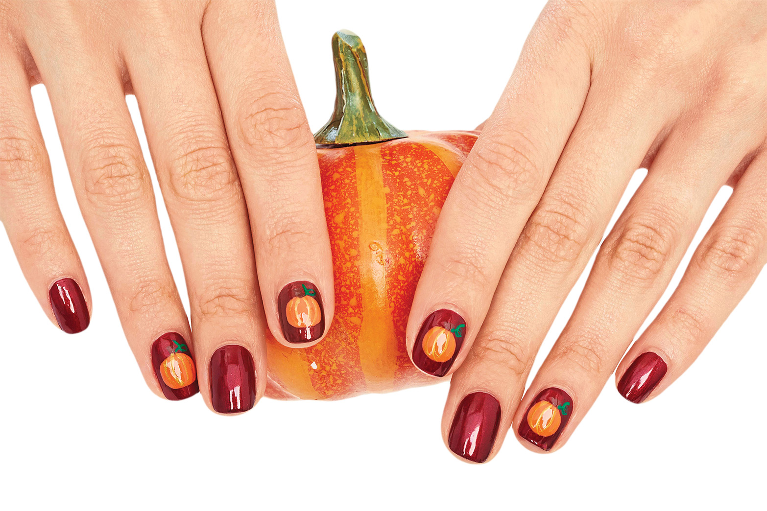 Hands holding a miniature pumpkin with nails painted dark maroon and with pumpkin fall nail designs accents