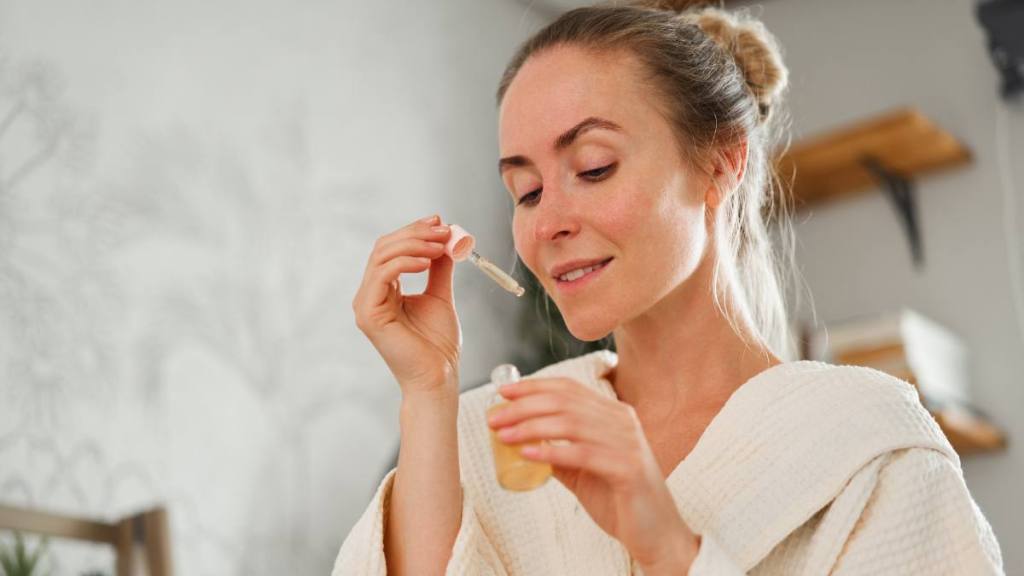 Work from home sales jobs: Smiling young female in bathrobe holding dropper and smelling aromatic essential oil while enjoying skincare session at home