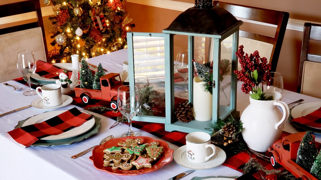 Christmas centerpiece ideas: Farmhouse centerpiece on dining table complete with a buffalo plaid runner, mini red pickup trucks, a pitcher of berry branches, a teal candle lantern and pine sprigs