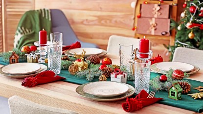 Christmas centerpiece ideas: Festive table setting for Christmas dinner featuring a satin green runner layered with candlesticks, evergreen boughs, gilded ornaments and mini Christmas village houses