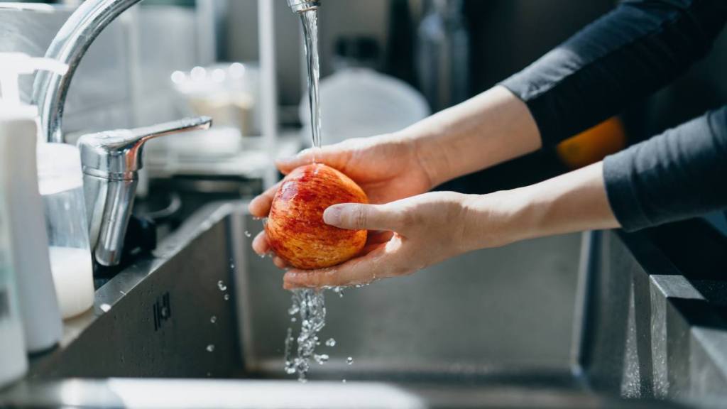 How to Clean Fruit: Cropped shot of woman's hand washing an red apple with running water in the kitchen sink at home