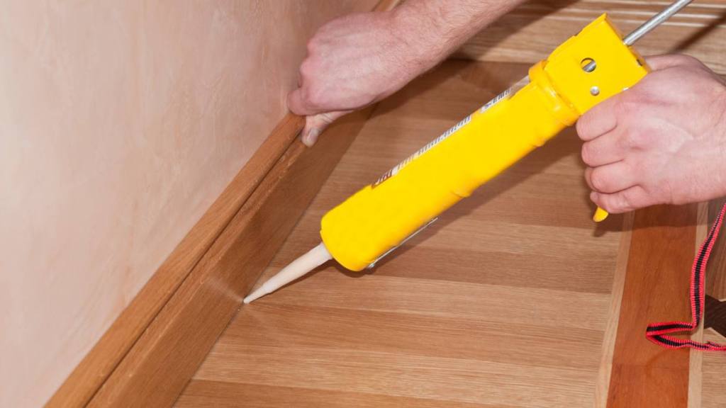 How to Silence a Squeaky Floor: Oak Wood floor installation. Carpenter on work putting wood parquet skirting board with glue. Caulking Gun in Hand.