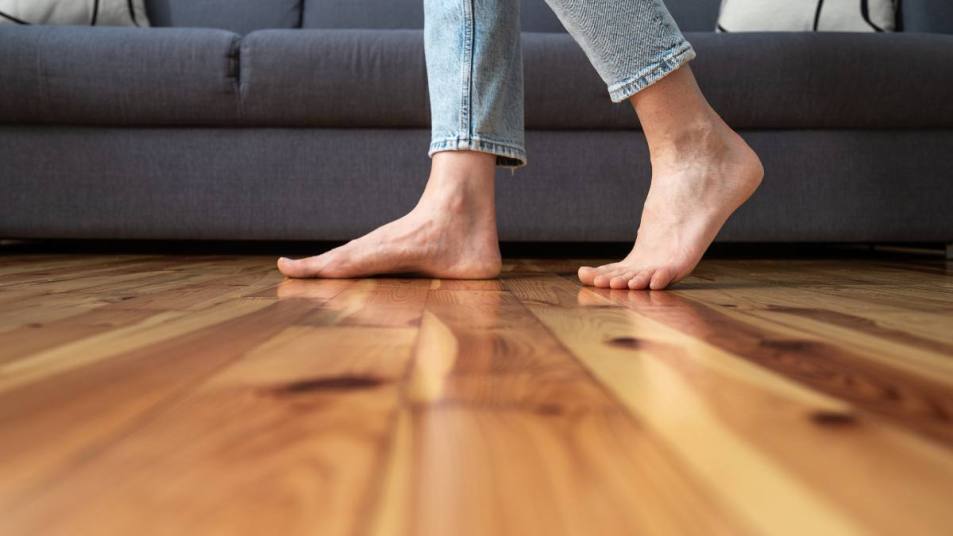 How to Silence a Squeaky Floor: cropped shot of female legs walk barefoot on wooden warm floor near couch in living room at home, heating concept