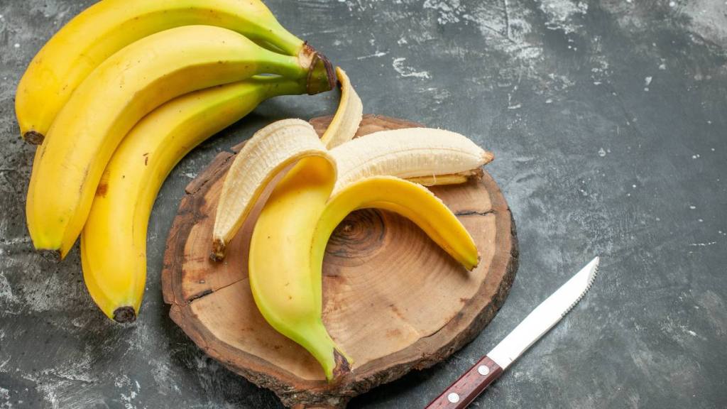 The best foods to eat for gut health: Top view nutrition source fresh bananas bundle and peeled on wooden cutting board knife on gray background