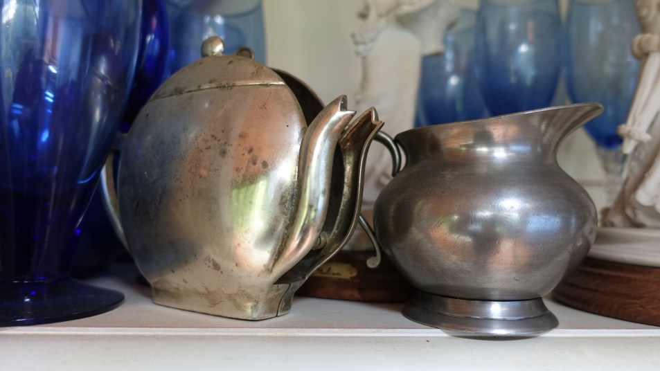 How to clean pewter: Pewter teapot and pitcher showing some oxidation on a shelf next to blue wine goblets and glasses