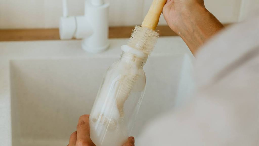 how to clean reusable water bottle: Close-up of young woman washing reusable glass bottle with natural brush at the kitchen