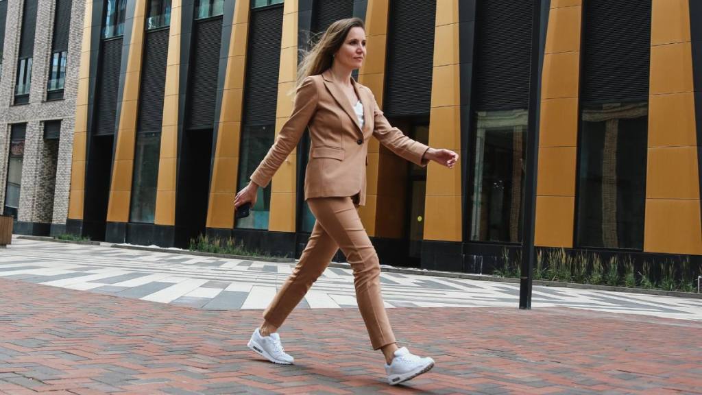 how to reduce stress at work: Business Lifestyle. Mid Adult Confidence Female Manager Wearing Beige Pant Suit Walking Fast Against Modern Office Background, Holding Smartphone, Smiling And Looking Away. Front View, Full Length, Ambient Light, Copy Space