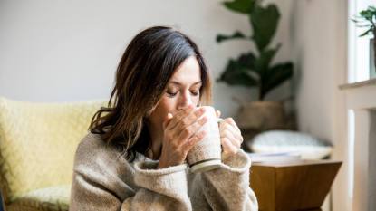 Woman with dark hair wearing a sweater sitting on a couch drinking a cup of tea for headaches