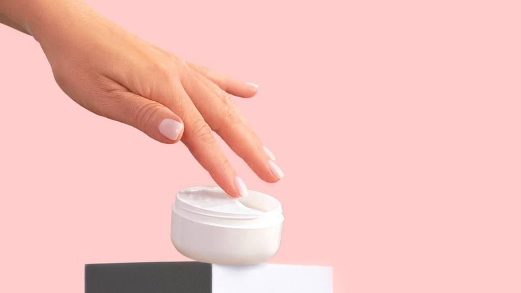 A close up of a woman dipping her hand into a jar of white cream with a pink background, which helps with menopause self-care
