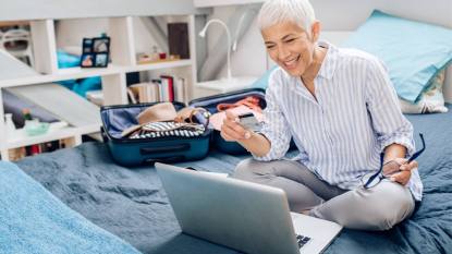 How to save money on travel: Mature woman packing her suitcase and using a laptop.