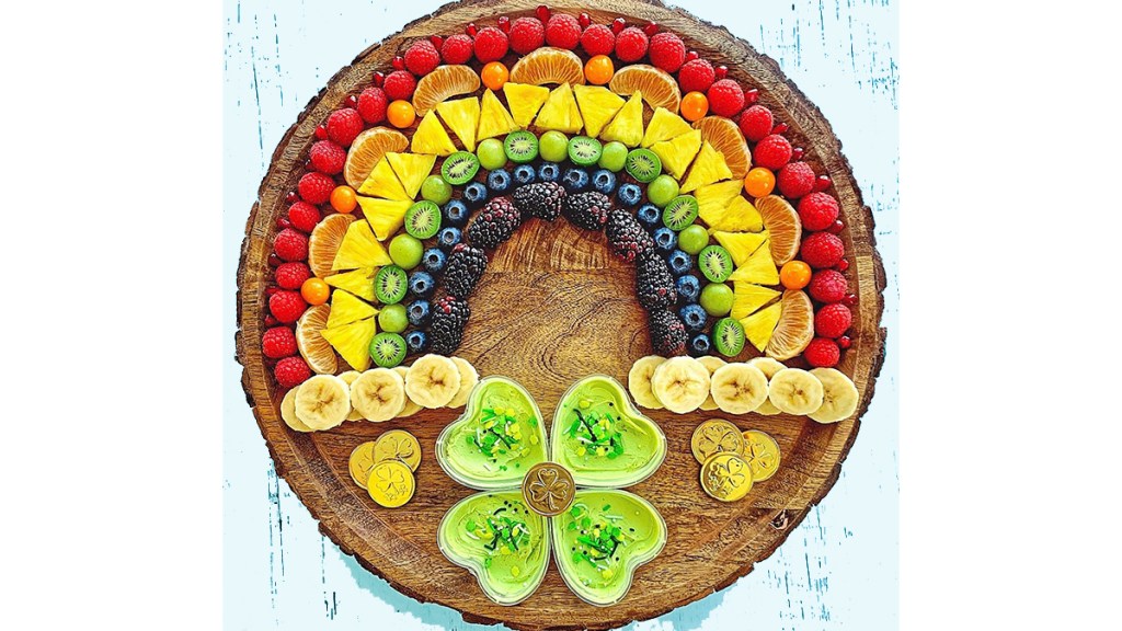 Snacks for St Patrick's Day: Sliced fruit and berries arranged on a round cutting board to look like a rainbow with sliced banana clouds, chocolate coins and a shamrock bowl filled with green fruit dip below it