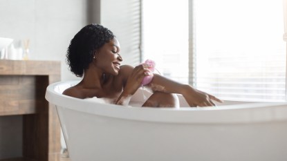 Side view of relaxed african american naked young woman taking bath at home, laying in beautiful bathtub, rubbing her skin with sponge and smiling, copy space. Home spa, hygiene, relaxation concept