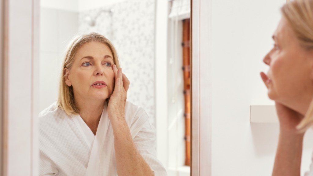 A woman with rosacea touching her face while looking in the mirror and wearing a white robe