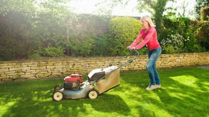 How to sharpen lawn mower blades: Mature woman mowing sunlit garden lawn with lawn mower