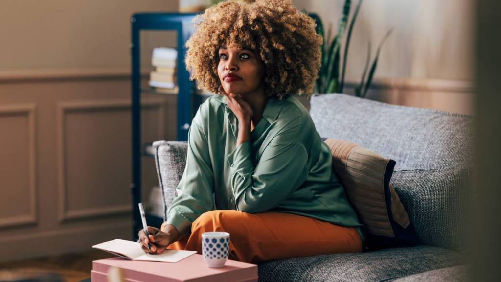  Pensive young African American woman sitting on a couch, jotting down notes in a notebook, surrounded by cozy home decor.
