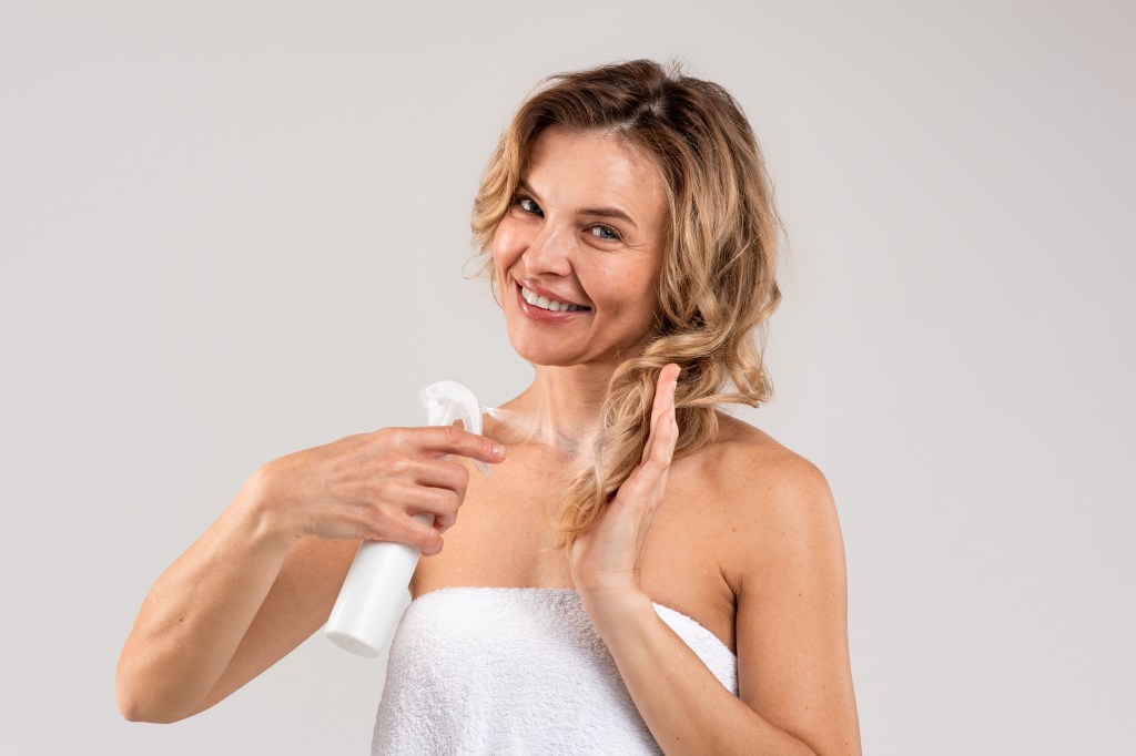 Mature woman spraying oil onto hair smiling.