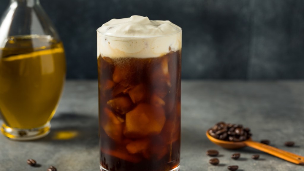 A glass of olive oil coffee topped with foam. Coffee beans are scattered around the glass and there is a bottle of olive oil in the background.