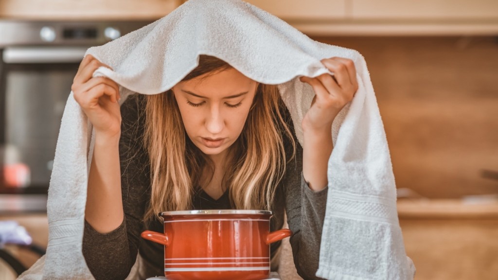 A woman with a towel over her head leaning over a pot of essential oils for congestion
