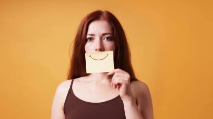 woman looking sad holding a post-it with a smiley face over her mouth: What is Smiling Depression?