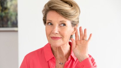 A woman in a coral shirt with her hand by her ear trying to hear better
