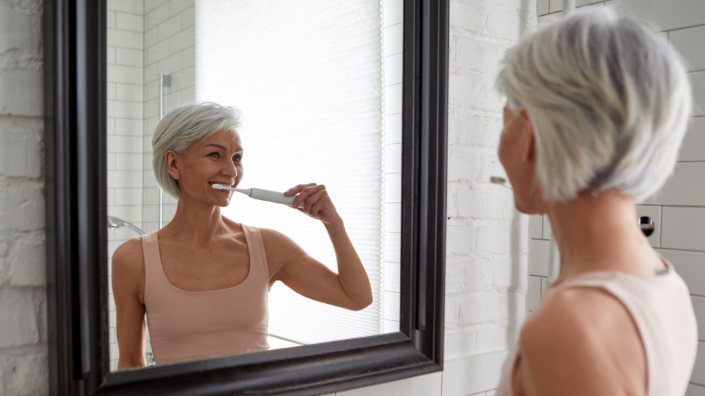A woman with short grey hair brushing her teeth in front of a mirror to eliminate bad breath from the stomach