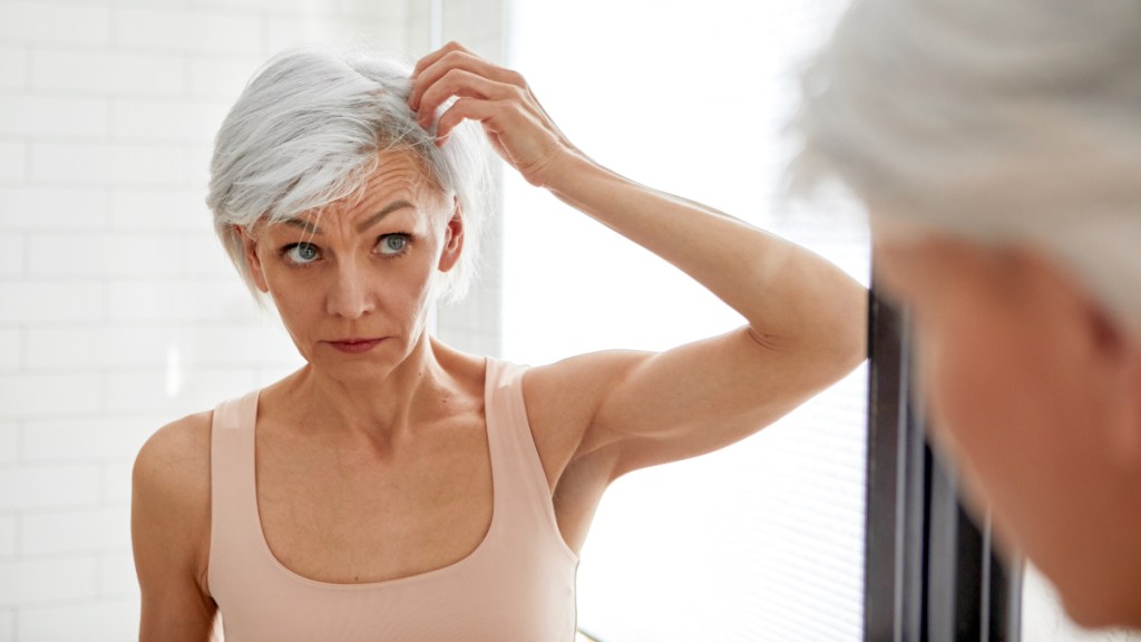 A woman in a tank top with short grey hair looking at her hair in a mirror