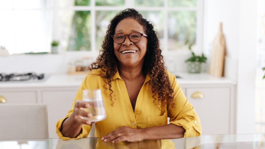 A woman wearing glasses and a yellow shirt who is holding a glass of water to help with bladder spasms