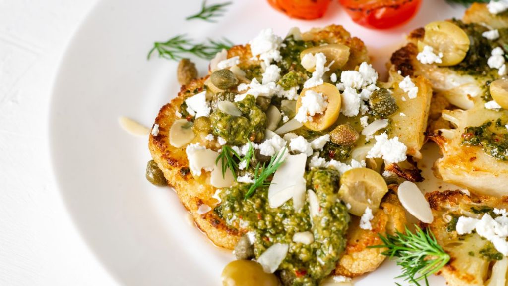 Cauliflower Steak with spices, chimichurri sauce, almond flakes, olives, fried cherry tomatoes and capers on a white plate