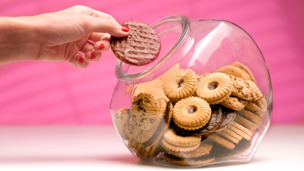 A close up of a woman with red nails reaching her hand into a cookie jar and taking a cookie