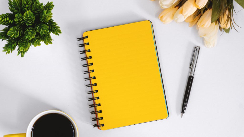A yellow journal on a white table with a cup of coffee, a plant, flowers and a pen