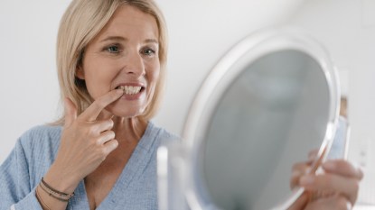 A woman with her finger on her tooth examining her white gums in a handheld mirror