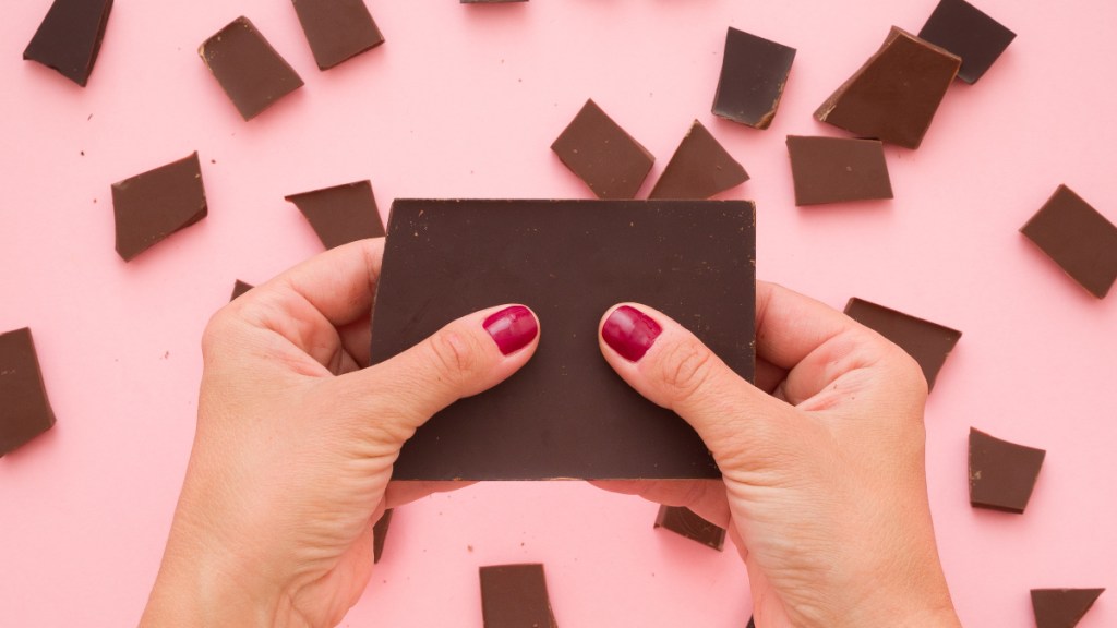A close-up of a woman's hands breaking a bark of chocolate over a pink background