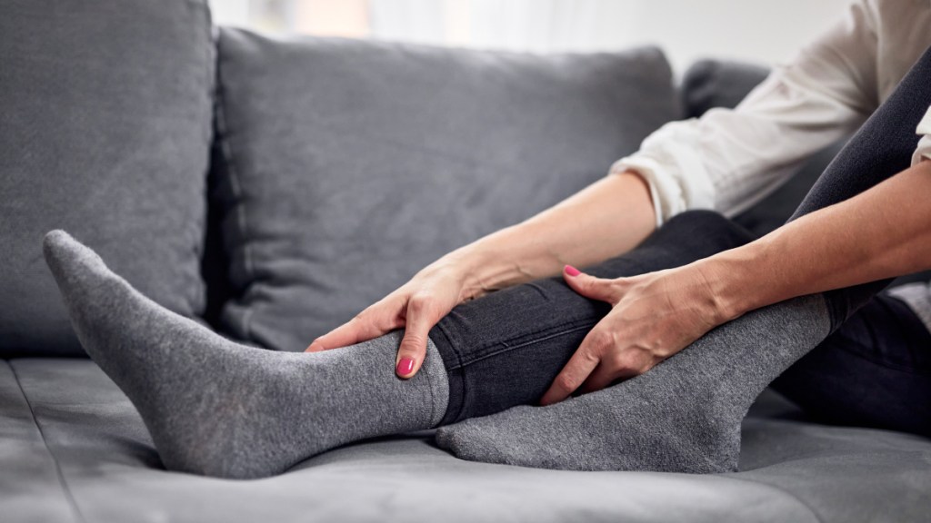 A close up of a woman with pink nails sitting on a couch holding her leg, which can be helped with physical exericise for restless leg syndrome