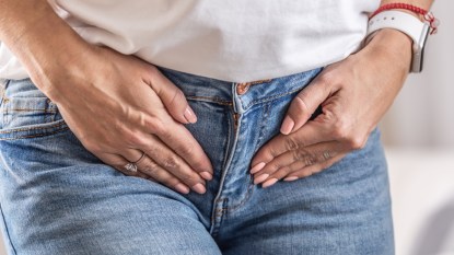 A close up of a woman in a white t-shirt and jeans holding her hands to her pelvic region due to hypertonic pelvic floor pain