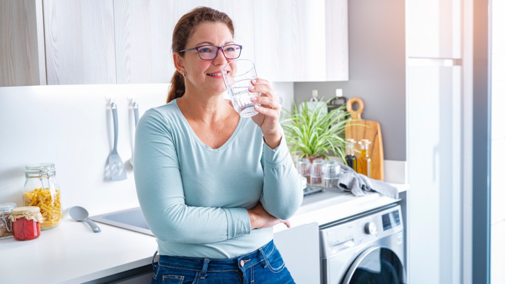 A woman wearing a blue shirt and jeans drinking a glass of water in her kitchen