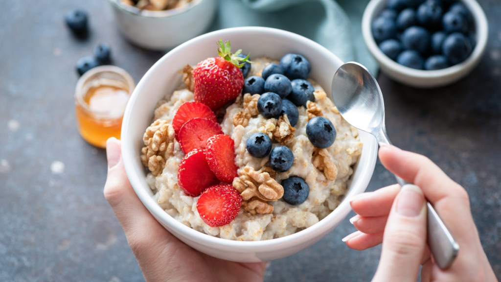 A woman's hands holding a bowl of oatmeal topped with nuts, strawberries and blueberries, which helps reduce mucus in urine