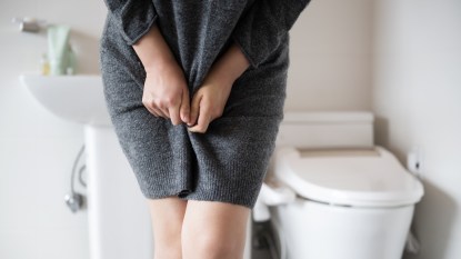 A close-up of a woman in a grey sweater dress holding her hands to her bladder due to excess mucus in urine while standing in front of a toilet