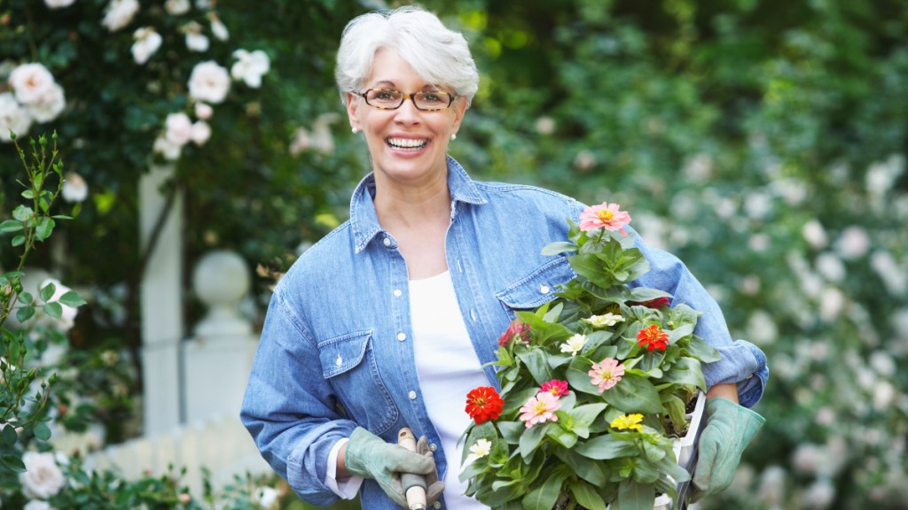 A woman with grey hair wearing a chambray shirt while holding flowers and a gardening shovel, which helps reduce mucus in urine