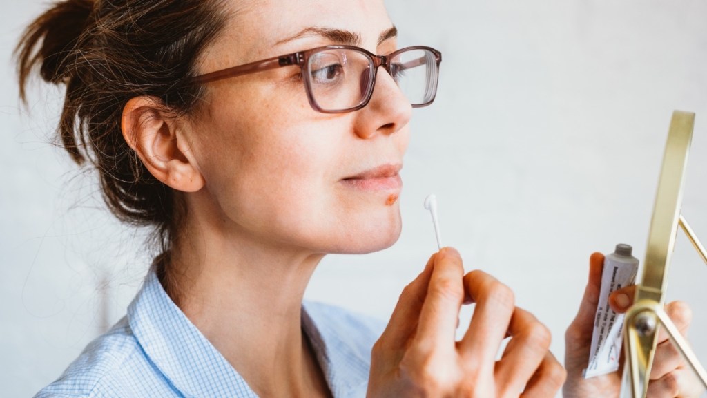 A woman wearing glasses while using a cotton swab to apply cold sore cream