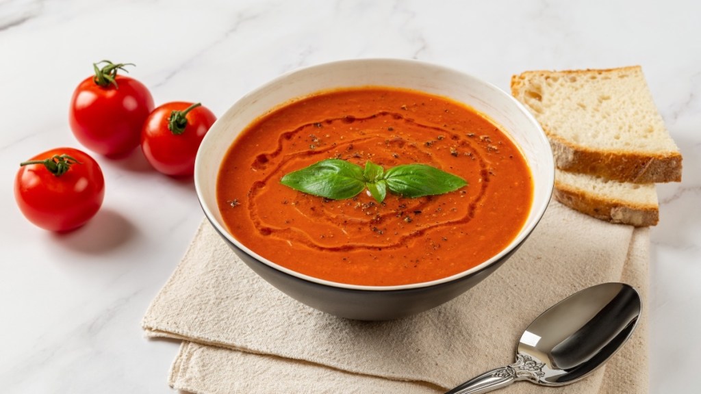 A bowl of tomato soup next to a spoon, fresh tomatoes and sliced bread