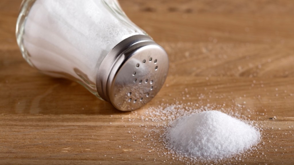 A salt shaker tipped over on a wooden table spilling salt