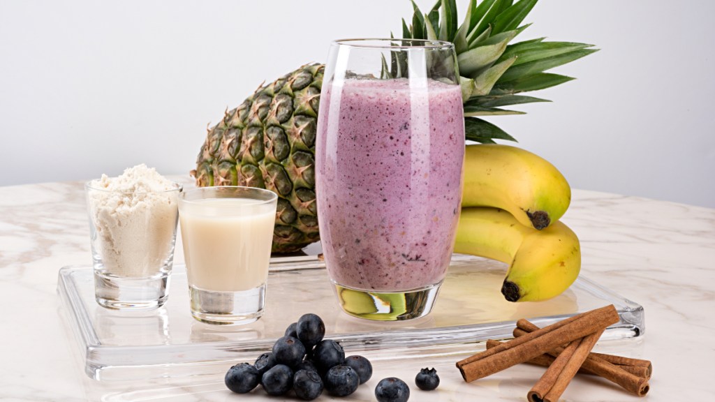 A blueberry smoothie beside protein, pineapple, banana and berries
