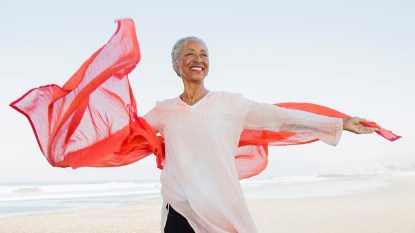 Happy mature woman on beach with outstretched arms and scarf blowing in wind