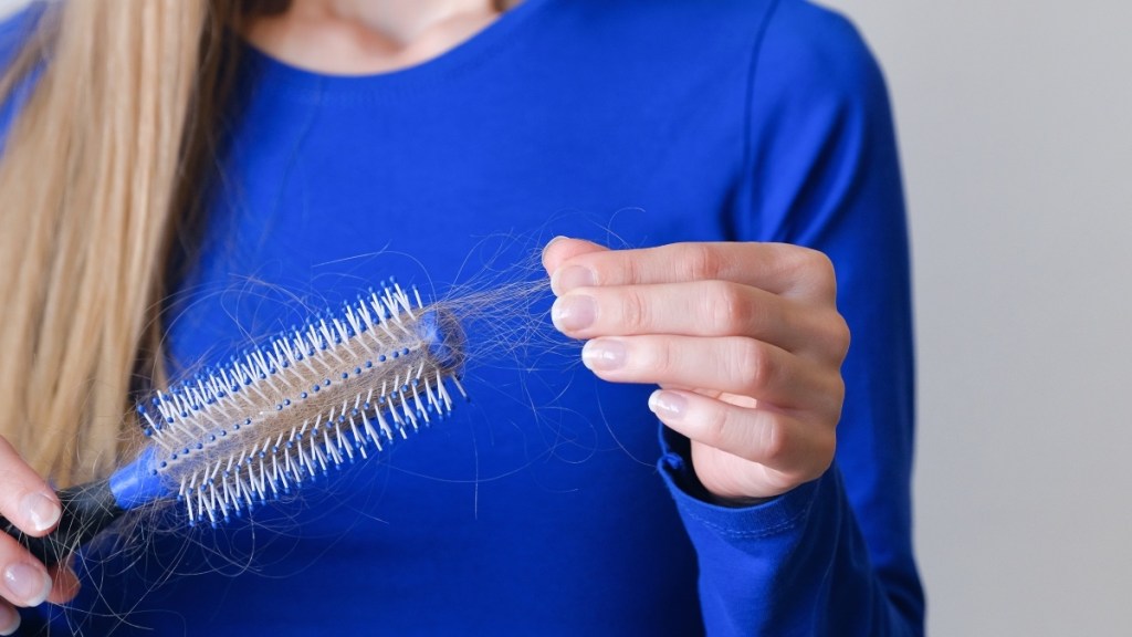 A close-up of a woman in a blue shirt pulling hair from a hair brush after dealing with hair loss