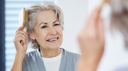 A mature woman brushing her hair while smiling in the mirror after using spironolactone for hair loss