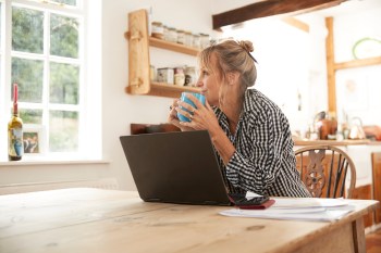 Smiling senior woman looking away while drinking coffee sitting with laptop in kitchen working from home home, walmart