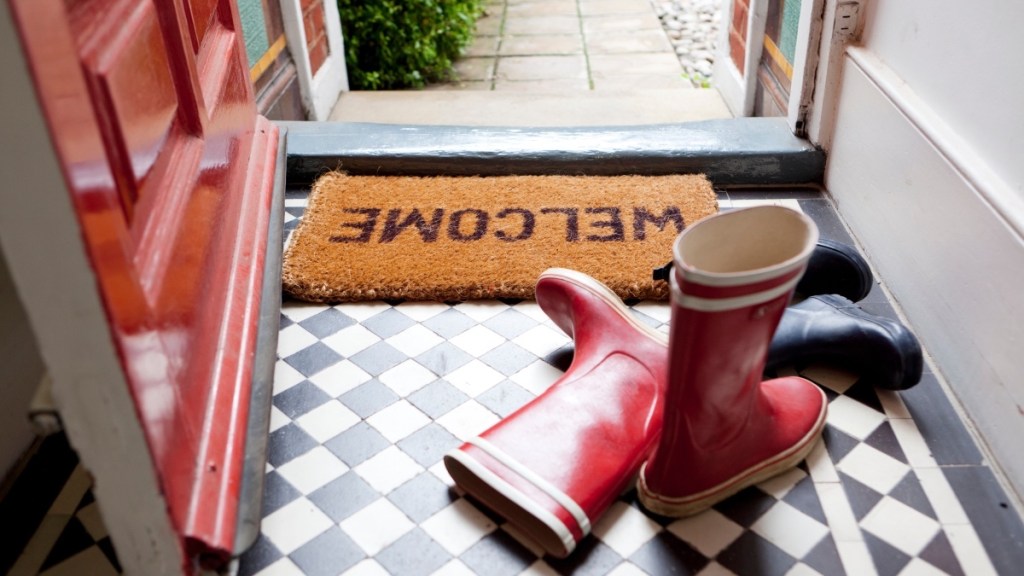 A black and white checkered floor with red boots by an open door and a welcome mat